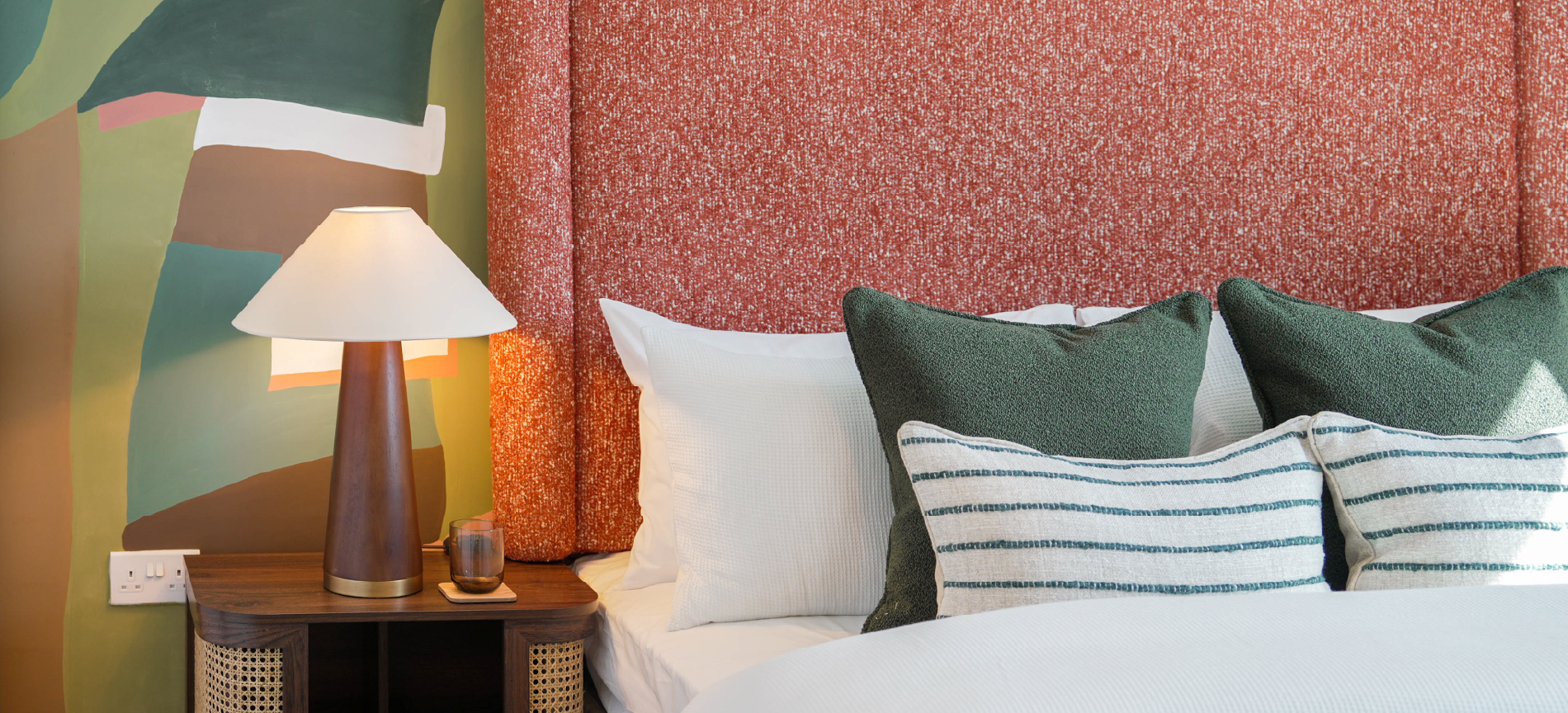 Cozy bedroom inside The Coanda Show Home, with a textured orange headboard, green and white striped pillows, a wooden side table, and a modern lamp. Earthy wall tones enhance warmth.