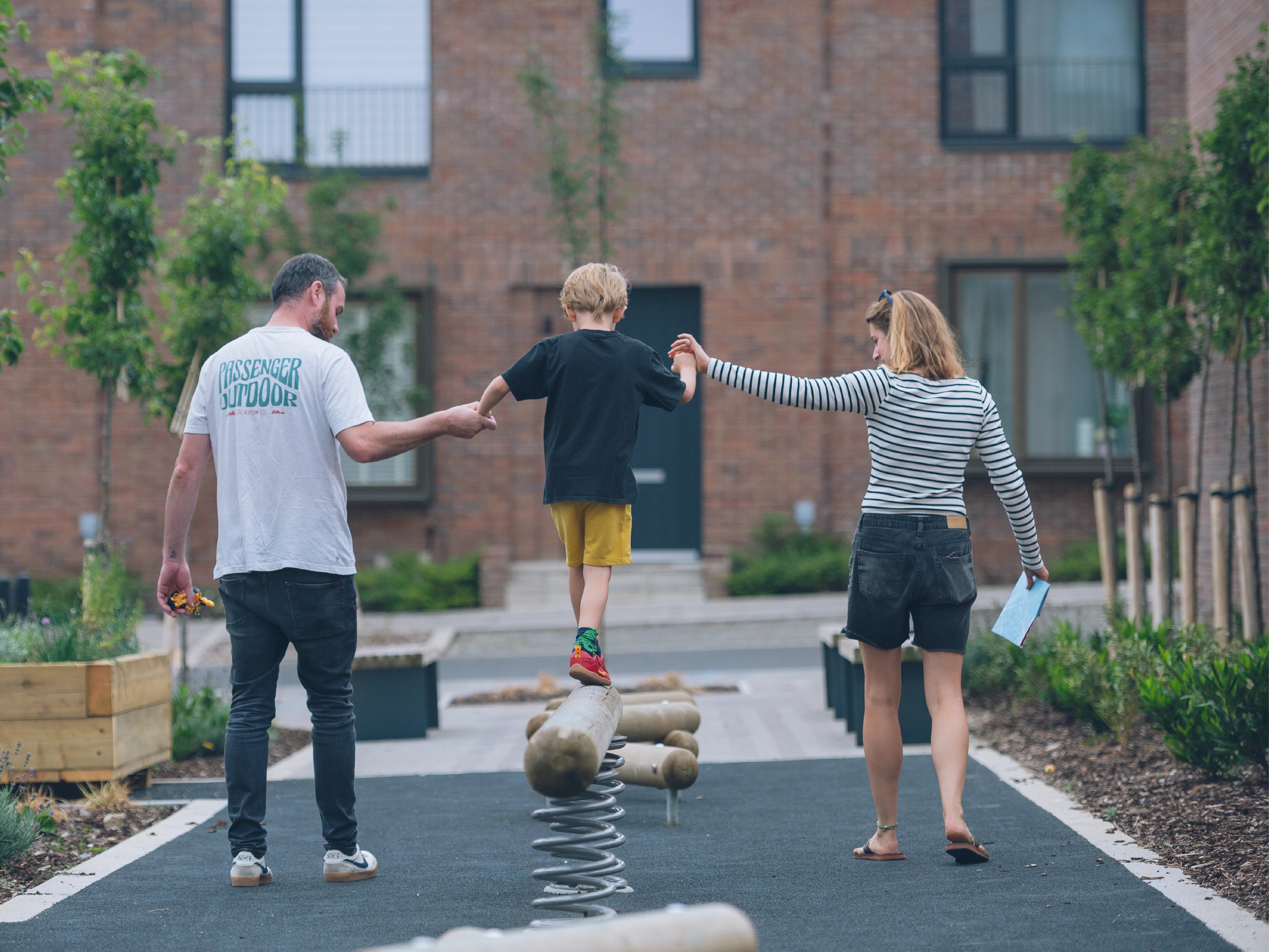 A child balances on a log spring in the pocket park at Brabazon, supported by two adults holding their hands. 