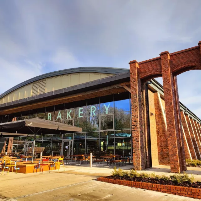 Exterior view of Spitfire Hangar - a thriving new community hub at Brabazon, North Bristol, showcasing a large 'Bakery' sign, outdoor seating and a window curtain.