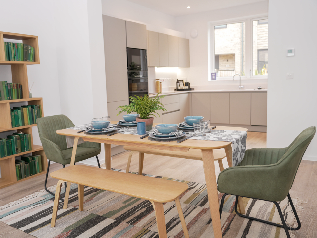 Modern dining area inside The Skylark, with a wooden table set, featuring green chairs, bench seating, and a light kitchen in the background.