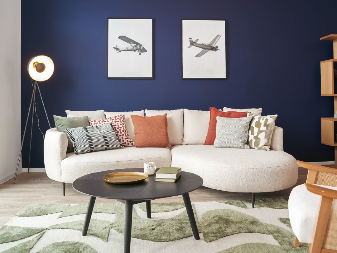 A modern living room with a curved white sofa, assorted patterned cushions, a round coffee table, and two airplane prints on a dark blue wall.