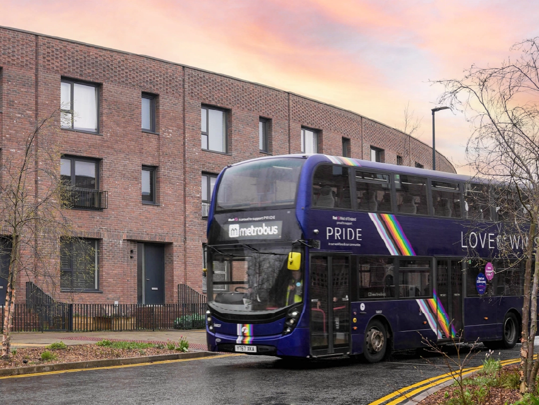 Deep blue First Metrobus with Pride stickers, driving down residential street at Brabazon on a rainy day.