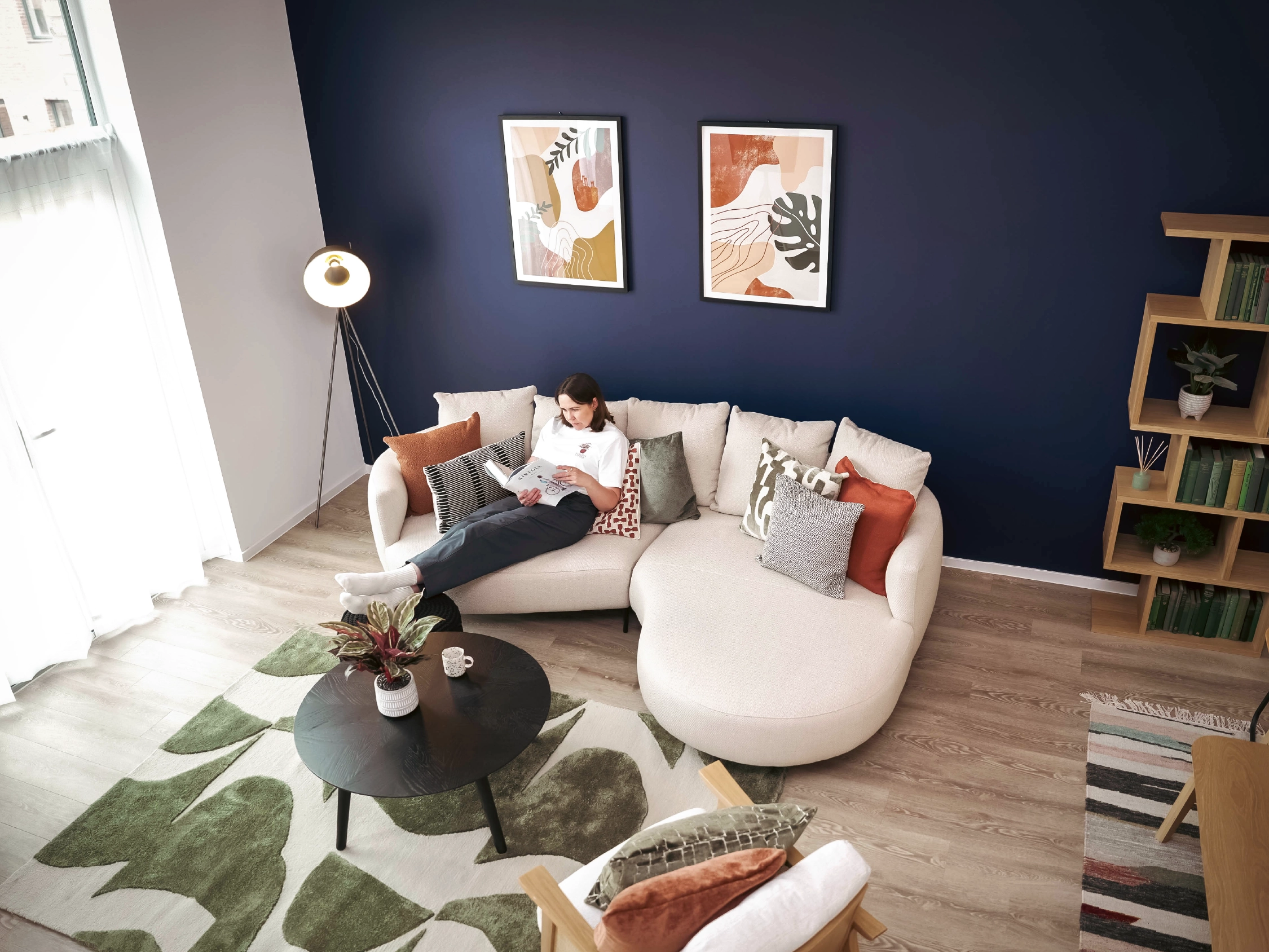 A woman reads on a curved beige sofa in a living room with a navy accent wall inside The Skylark living room. The living room features abstract artwork, a tripod floor lamp, and a round coffee table.
