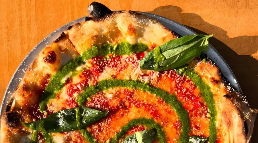 Close-up of a pizza topped with basil leaves, cheese and pesto on a table.