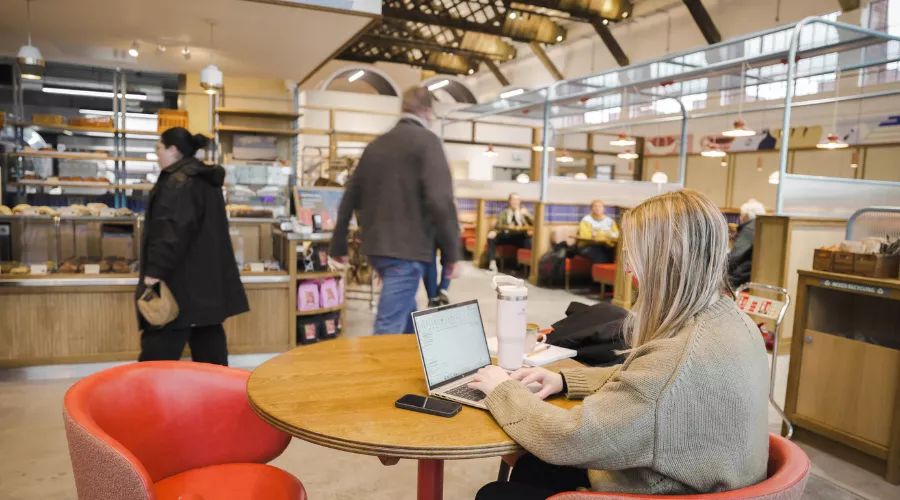 A woman sits at a round table inside Mokoko Bakery at Spitfire Hangar, typing on a laptop. Other customers are visible in the background near a pastry counter. 