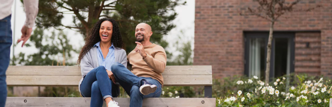  A man and woman sit on a park bench at Brabazon, smiling at someone just out of camera shot. 