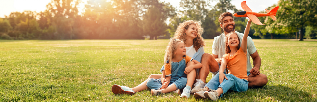 A joyful family of four sits on grass in a sunlit park. Two girls, one holding a toy plane, smile with their parents.