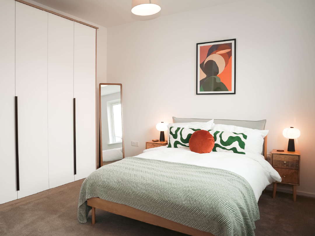 Principal Bedroom in The Skylark: a 3-bedroom home in bBistol. The room features large fitted wardrobes included as standard, a wooden bed with green and orange pillows, bedside tables, a floor mirror, and abstract wall art.
