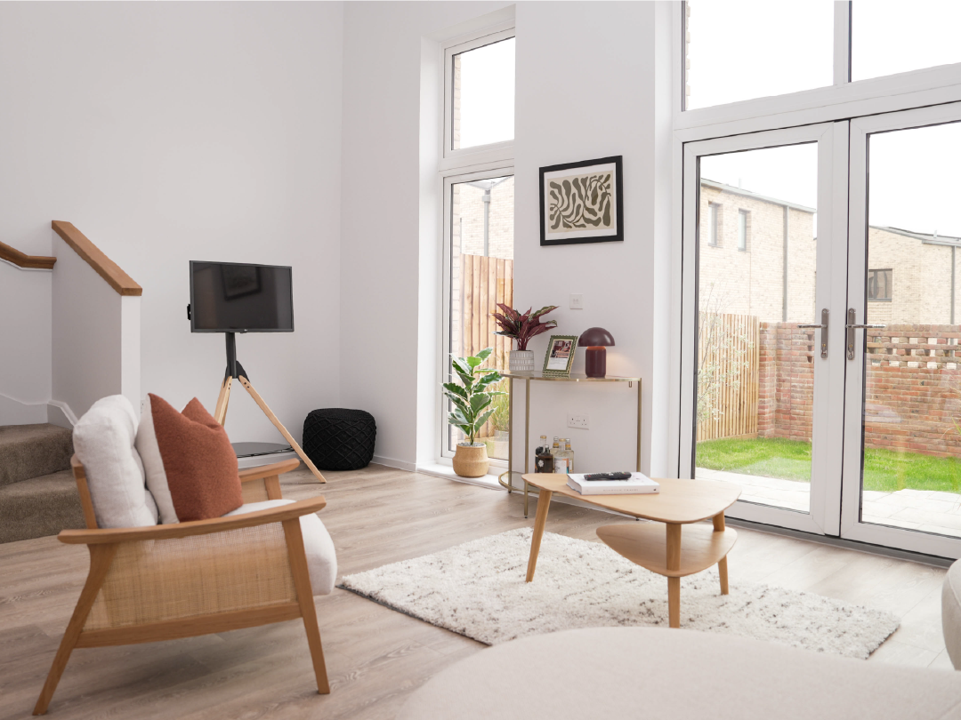 Modern living room inside The Skylark,  with tall windows, light wood furniture, and soft neutral tones. A cozy ambiance with natural light and a view of a green backyard.