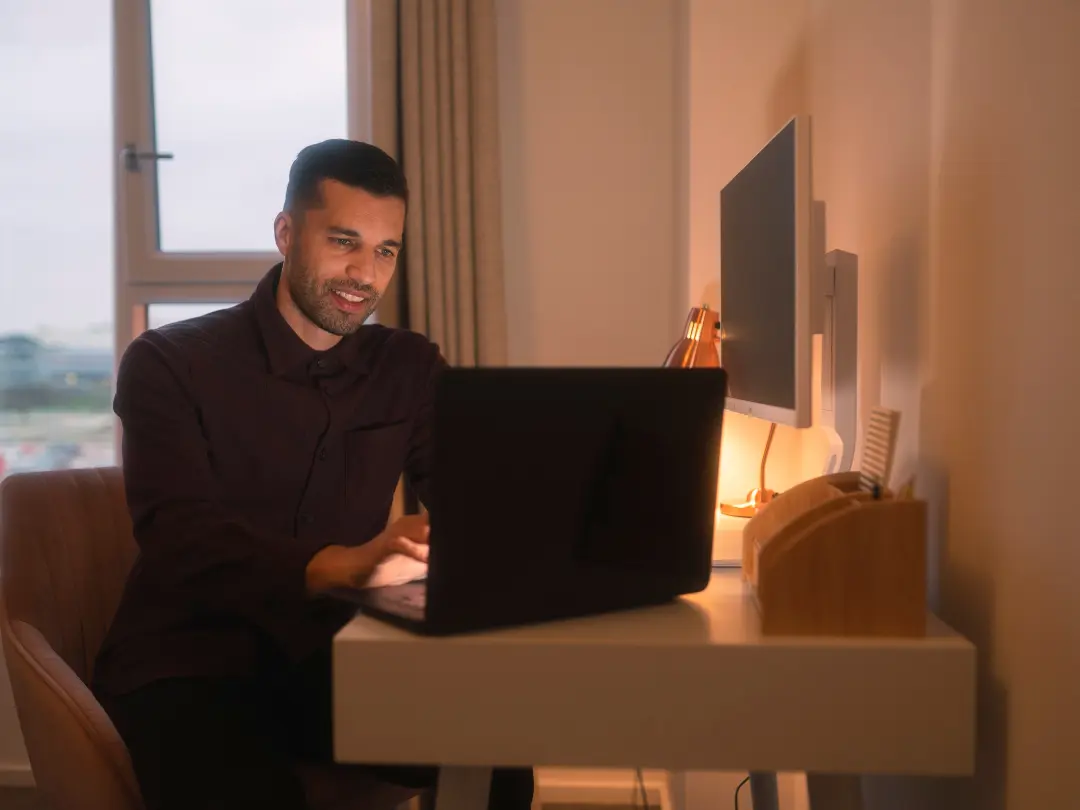 A man sits at a desk using a laptop in a warmly lit room, looking focused and relaxed. 
