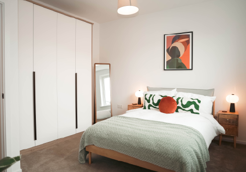 Principal bedroom inside The Skylark Show Home, featuring a double bed, green and white pillows, a round orange cushion, and a green throw blanket. Abstract art hangs above two bedside tables with lamps. 
