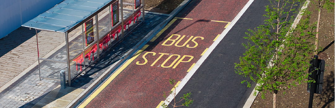 Aerial view of a Metrobus stop on Runway Avenue at Brabazon. The words "BUS STOP" are painted in yellow on the road. Nearby, there's a shelter with red benches, a clear roof, and surrounded by young green trees.
