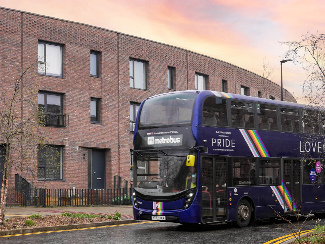 A double-decker bus with a Pride-themed design featuring rainbow stripes travels through Brabazon. 