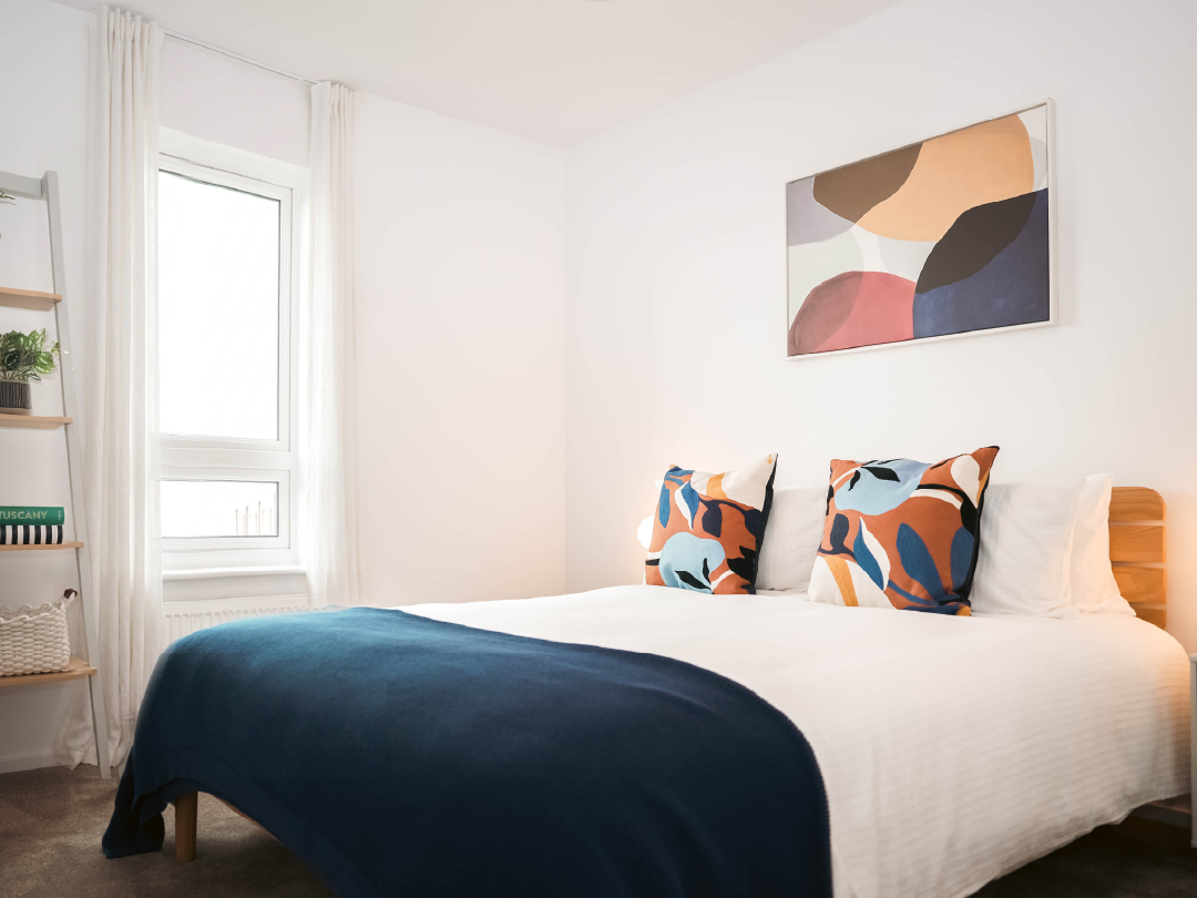 Bright second bedroom in The Skylark Show Home, feauturing a large bed, blue blanket, colourful throw cushions, abstract wall art, tall window with white curtains, and wooden shelving with plants and books.