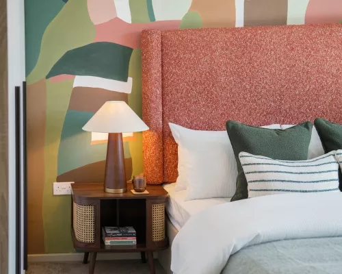A cozy bedroom inside The Coanda Show home, featuring a textured pink headboard, a bedside lamp, and colorful abstract wall art. Soft bedding and decorative pillows add warmth.