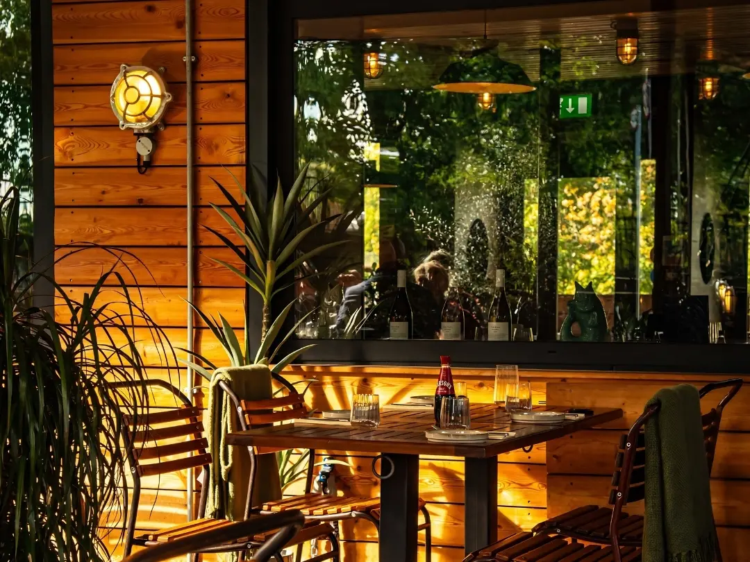 Sunlit outdoor restaurant terrace with wooden furniture, plotted plants and a table setting beside a large window.