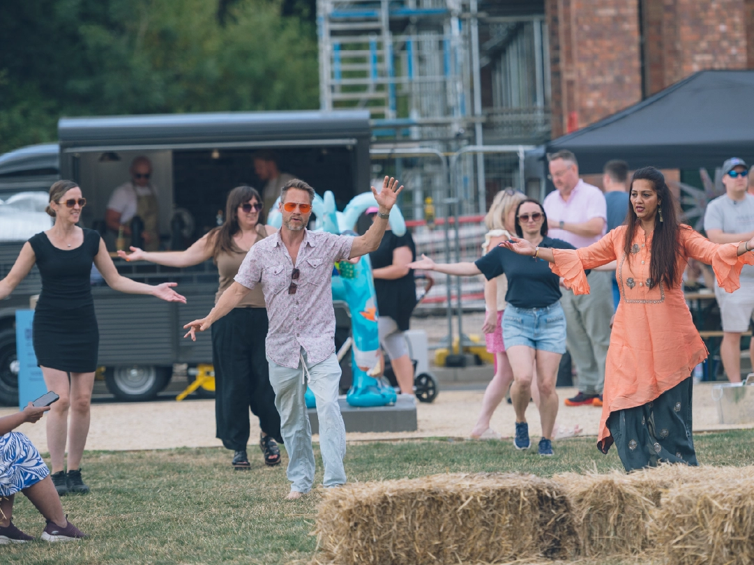 A group of people enjoy Bollywood dancing outdoors on The Green at Brabazon, Bristol. Behind sits a with a coffee cart. 