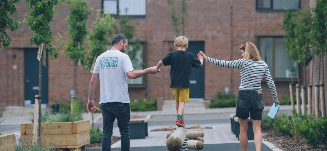A child balances on a climbing frame in the pocket park at Brabazon, holding hands with two adults.