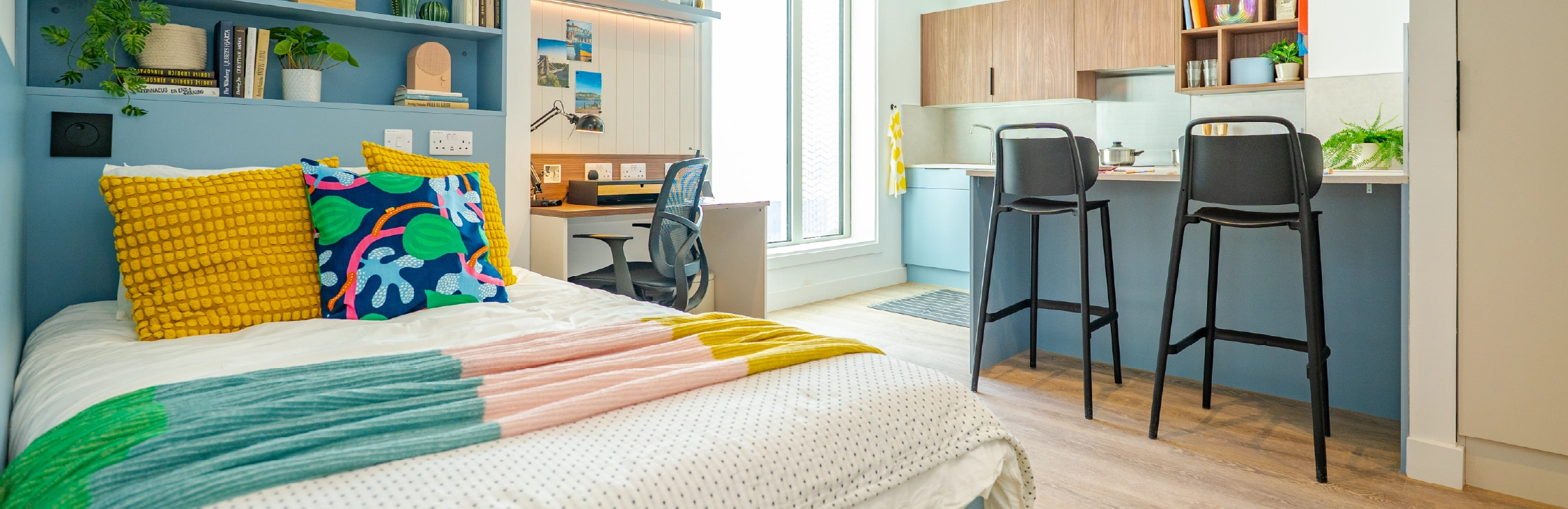Bright, modern studio flat inside The Propeller Quarter at Brabazon, with a neatly made bed adorned with colourful pillows and a throw. A desk with a chair is near a window, and a stylish kitchenette with two high chairs is in the background. 