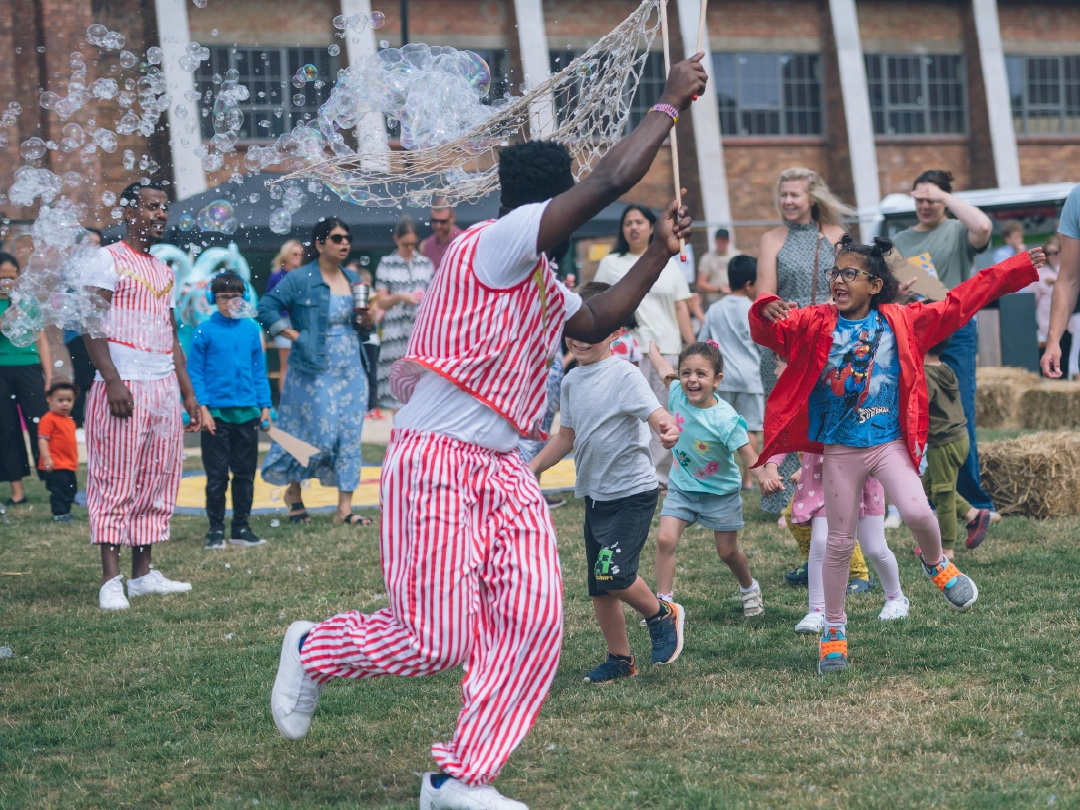 A cheerful outdoor festival at Brabazon, North Bristol. A performer in red and white stripes creating bubbles while excited children chase bubbles, while adults watch joyfully.
