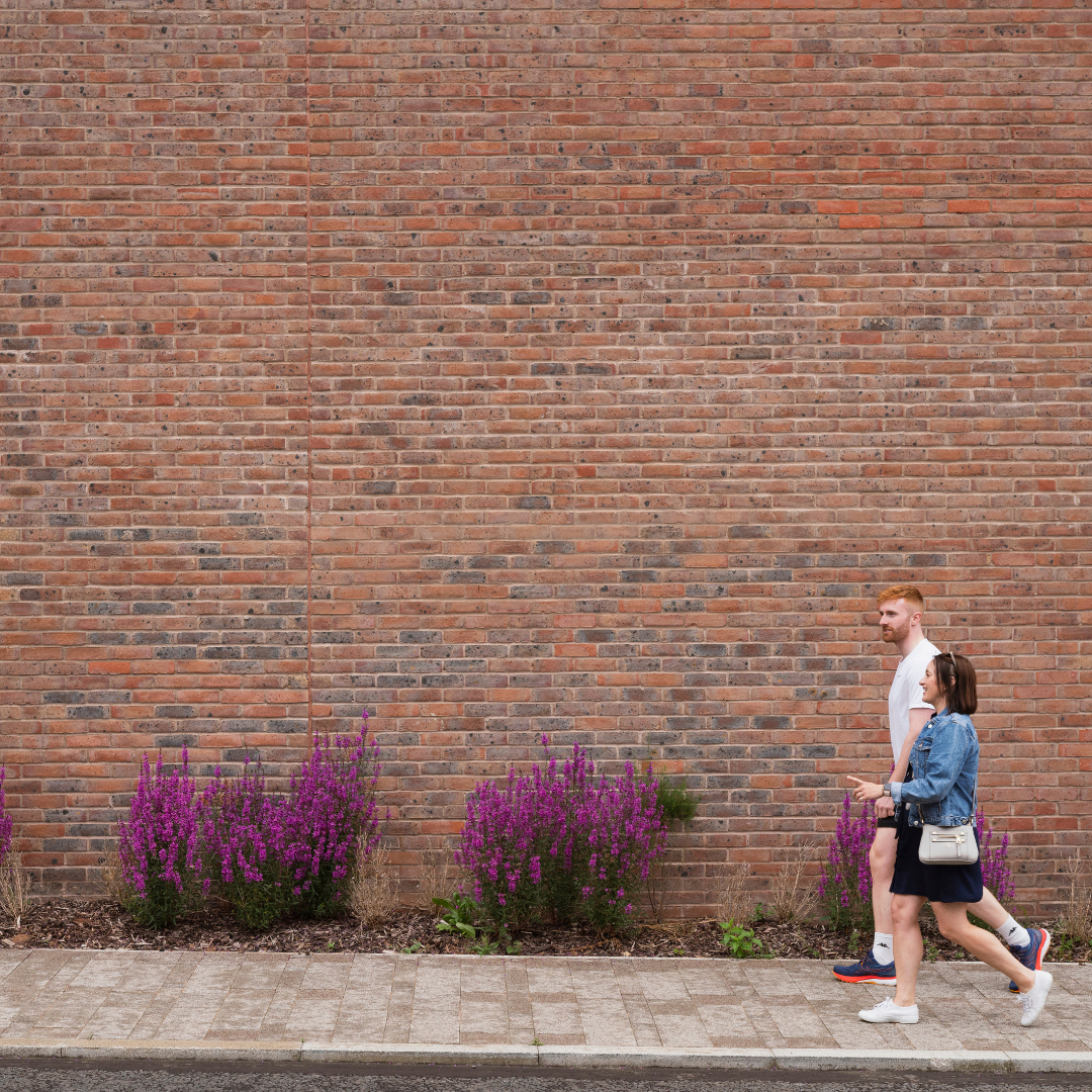 People in conversation walking along a street in front of a red brick wall.