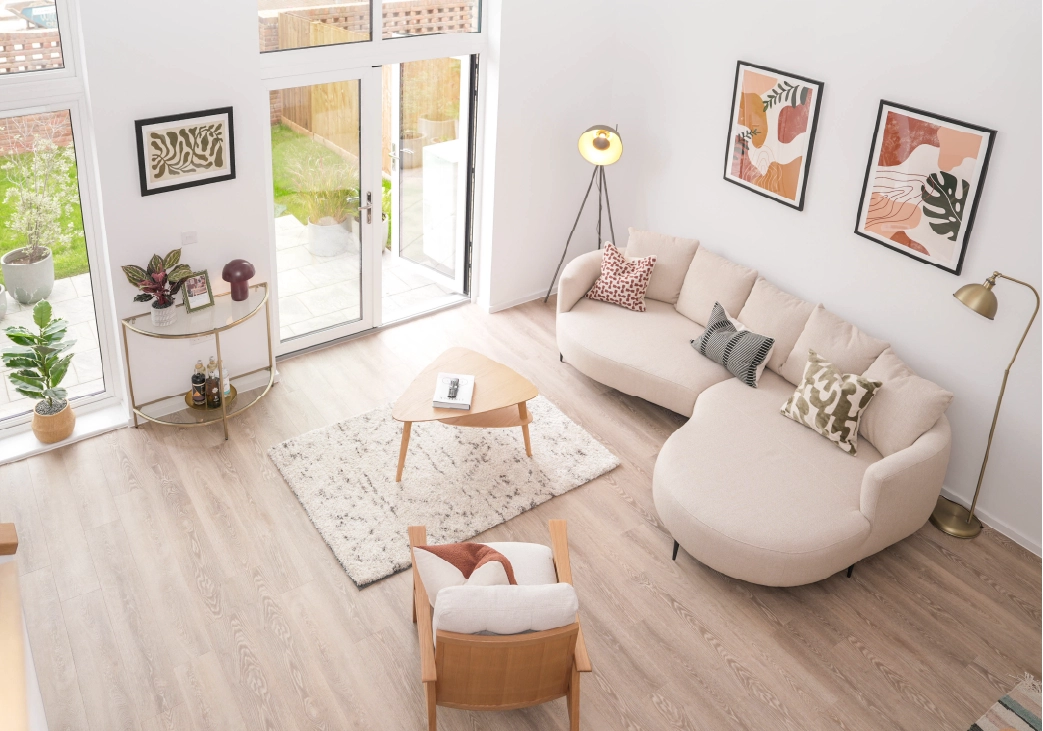 Modern living room inside The Skylark show home at Brabazon, with a light beige curved sofa, patterned cushions, and abstract art on white walls. Wood floors, floor lamp, and lush plants. 