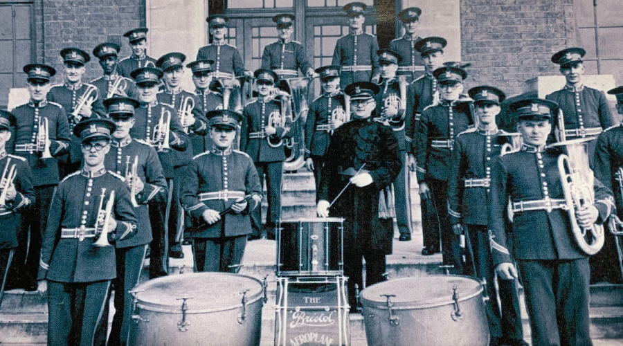 The men in the BAC brass band in 1940 dressed in uniform and holding instruments on the steps of a building.