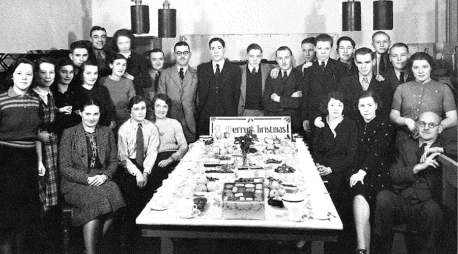 A large group of smiling men and women sat around a table of hot drinks and cakes in business attire.