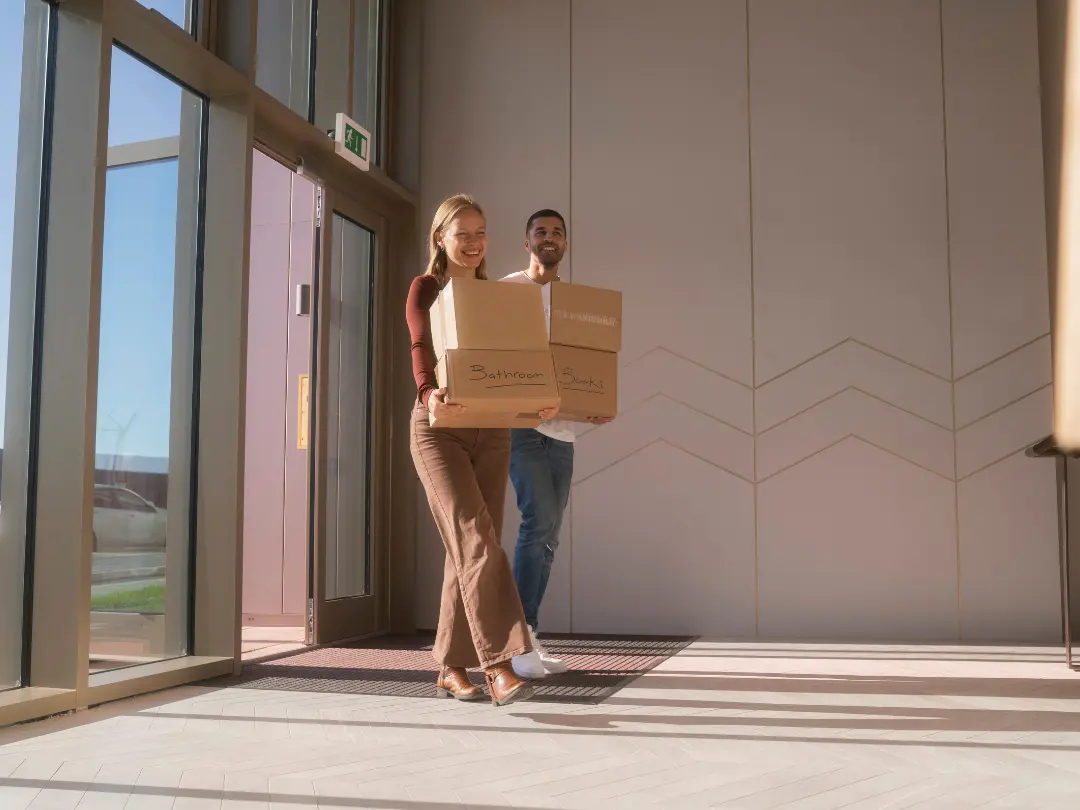 A smiling couple carries labeled moving boxes into a sunlit modern home at Brabazon.