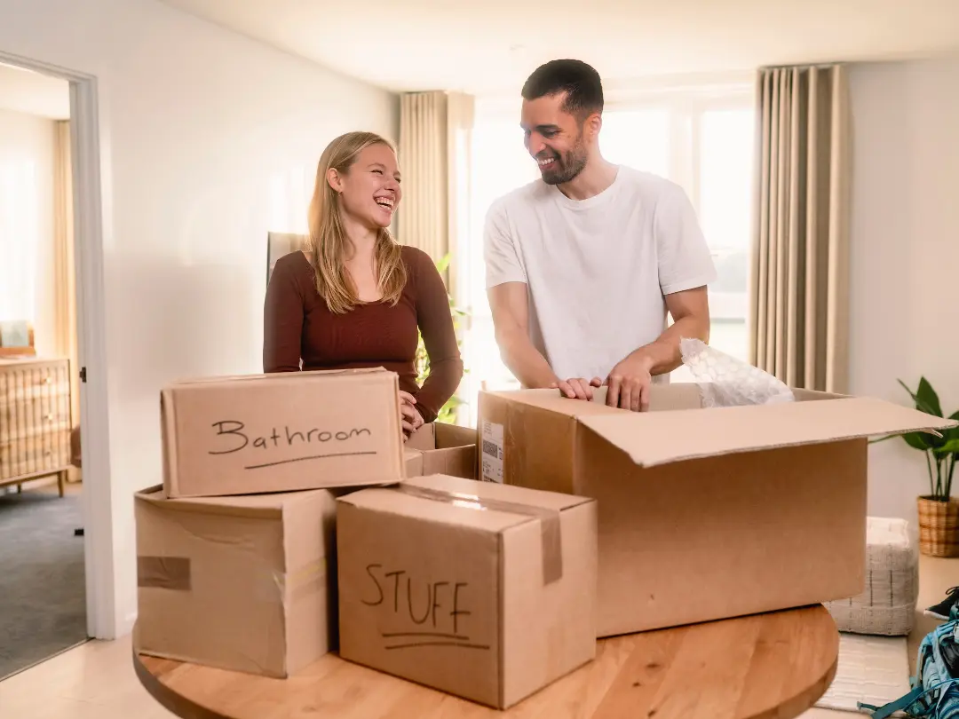 A smiling couple stands in a sunlit room surrounded by labeled moving boxes, suggesting a joyful packing experience.