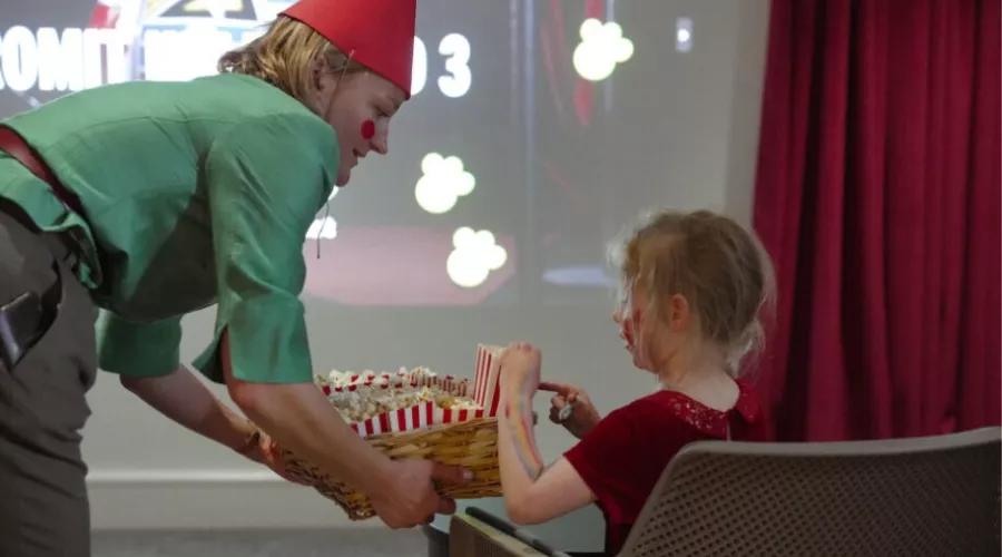 A person dressed as a gnome with a red hat and green shirt hands popcorn to a seated child in a theatre.