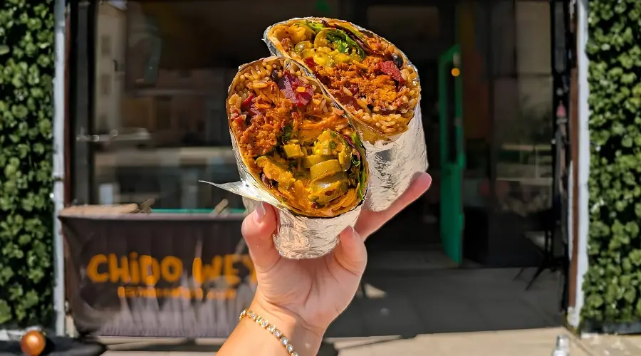 Hand holding a foil-wrapped burrito in two halves, filled with rice, vegetables and toppings, outside of a restaurant.