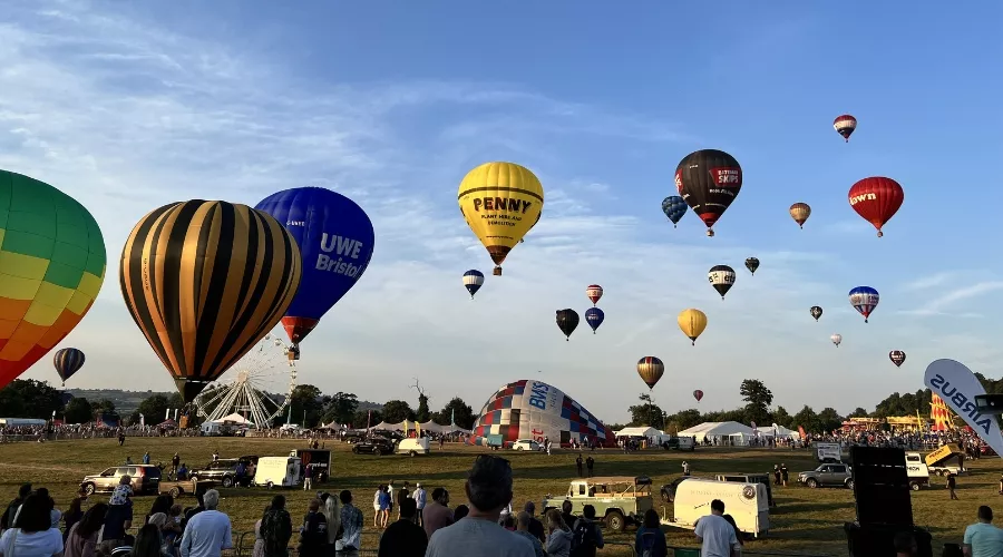 Hot air balloons of various colours and designs ascending at Bristol International Balloon Fiesta, with crowds and tents under a clear sky.