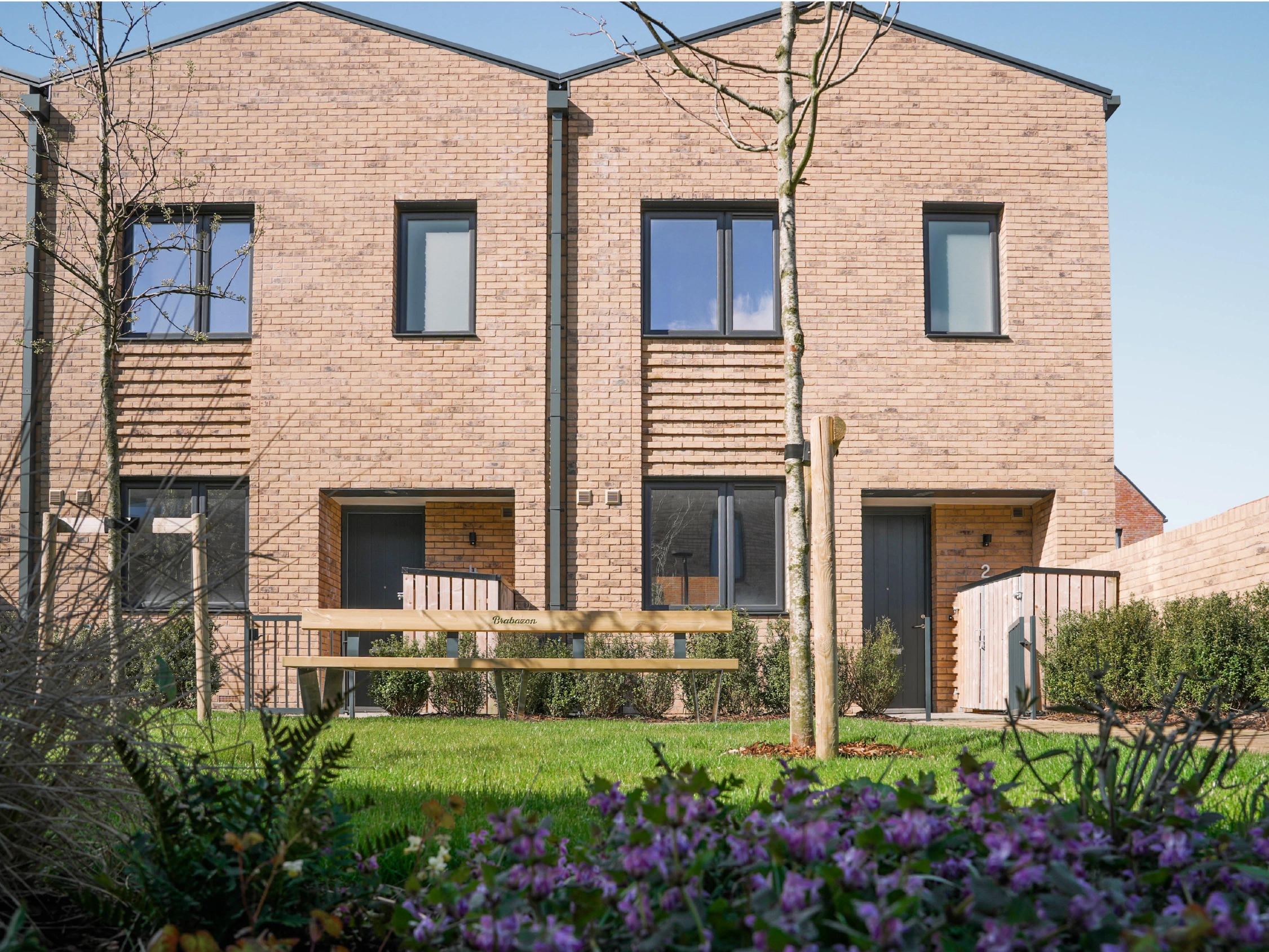 The Skylark house type at Brabazon.Two modern brick townhouses, featuring large windows and wooden doors. A bench, a tree, and purple flowers are in the foreground. 