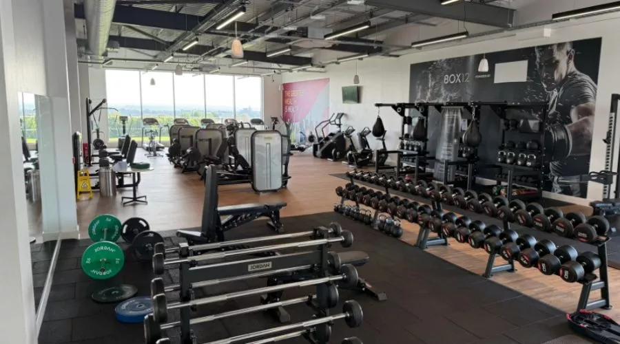 Spacious gym at Elm Park Leisure Centre, with weights, barbells, and cardio machines. Bright natural light filters through large windows. 