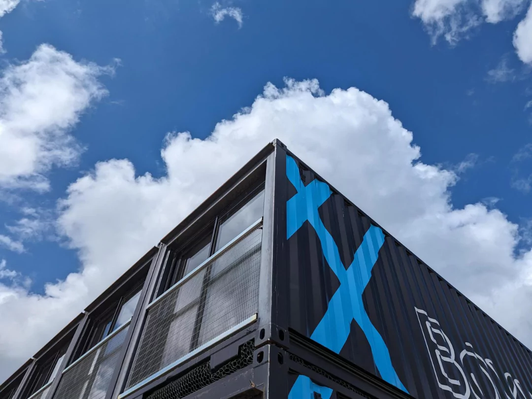 Boxworks' modern black shipping container offices stand against a bright blue sky with clouds.