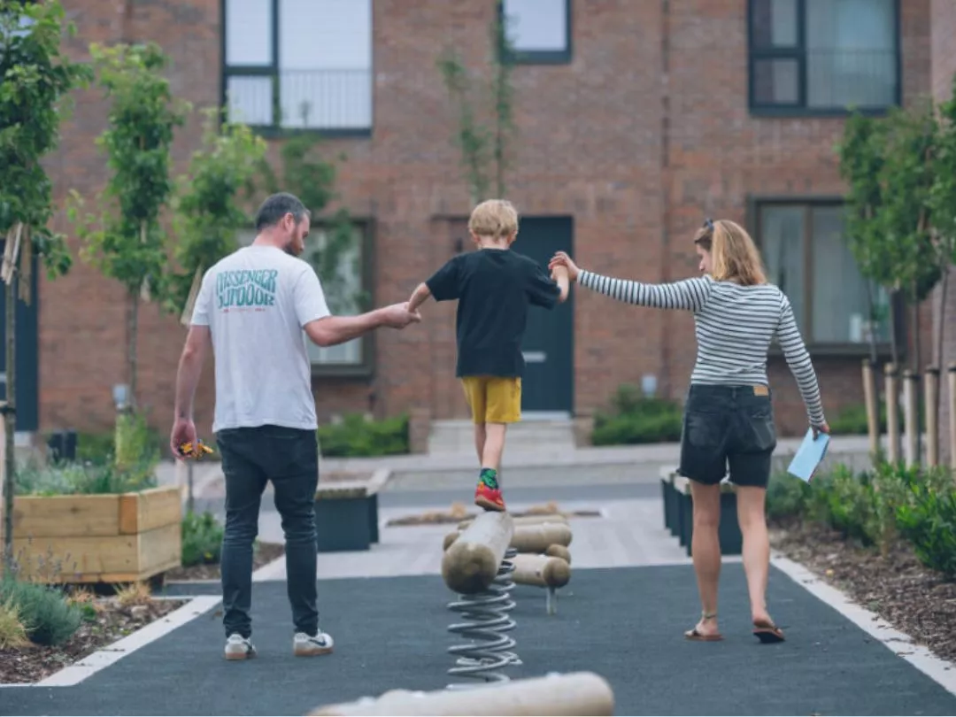 A child balances on a springboard holding hands with two adults in the pocket park at Brabazon, Bristol. 